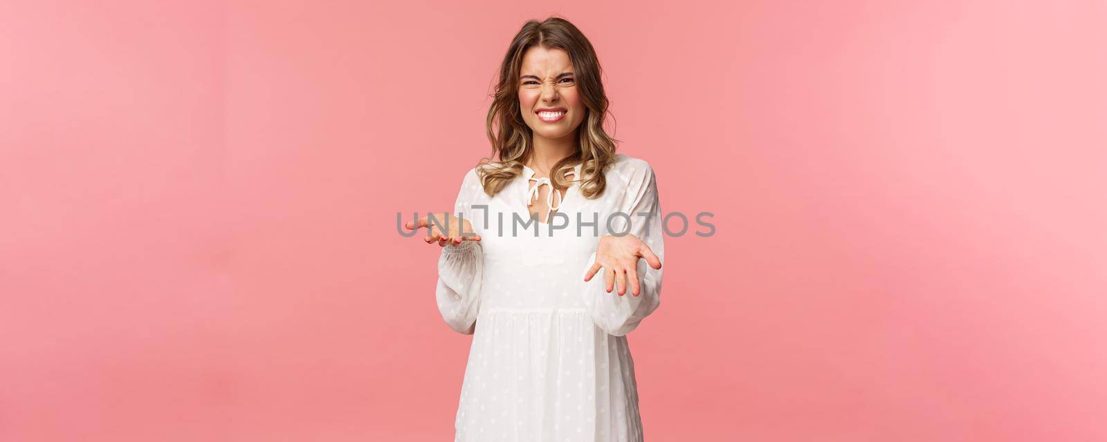 Portrait of dissatisfied, disappointed cute girl in white dress, cringe show tongue, smell something awful, pointing with raised hands at bad unappropriate thing, grimacing dislike, pink background.