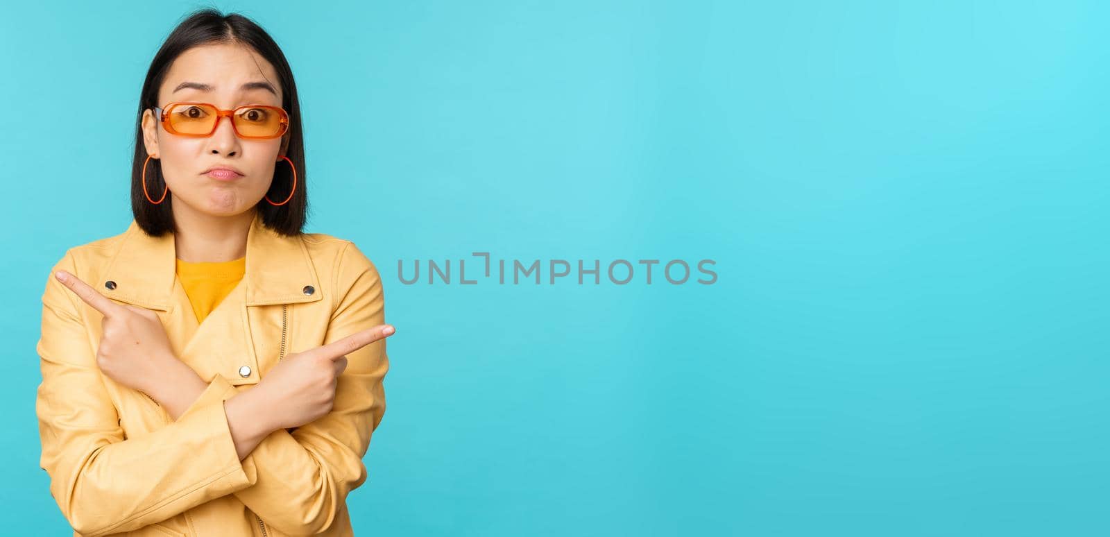 Indecisive asian woman in sunglasses, makes choice between two variants, points sideways, looks puzzled, stands over blue background.