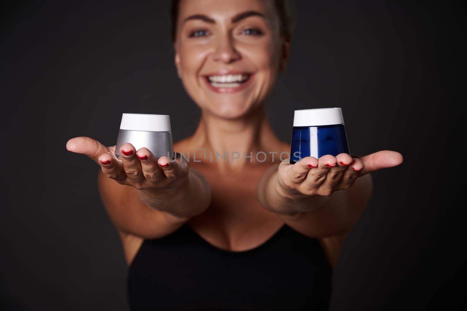 Focus on anti wrinkles moisturizer jars in the hands of blurred smiling middle aged blonde woman with tannes healthy clean skin, isolated over black background with copy ad space. wellness spa concept