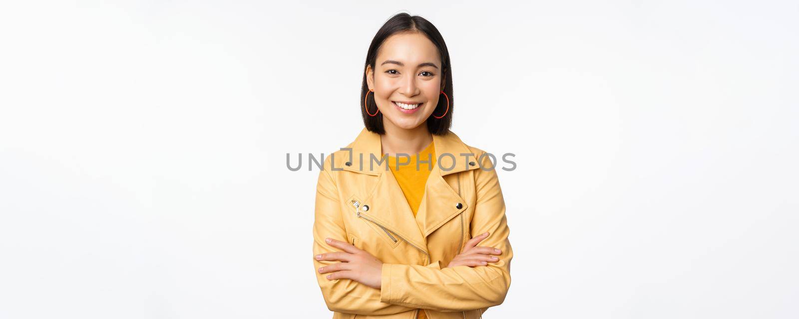 Portrait of asian woman in yellow jacket, smiling and looking happy, standing over white background. Copy space