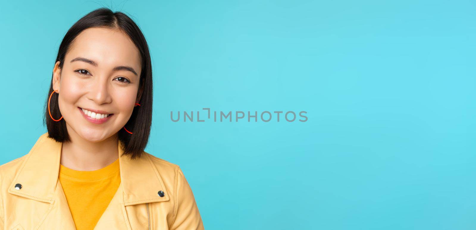 Close up portrait of smiling beautiful asian woman with white teeth, looking happy at camera, posing in yellow jacket over blue studio background.