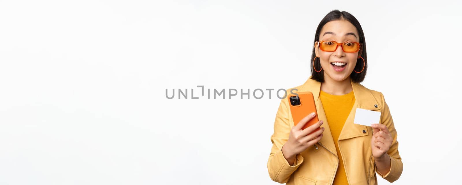 Online shopping. Stylish asian female model in sunglasses, holding credit card and mobile phone, smiling happy, standing over white background. Copy space