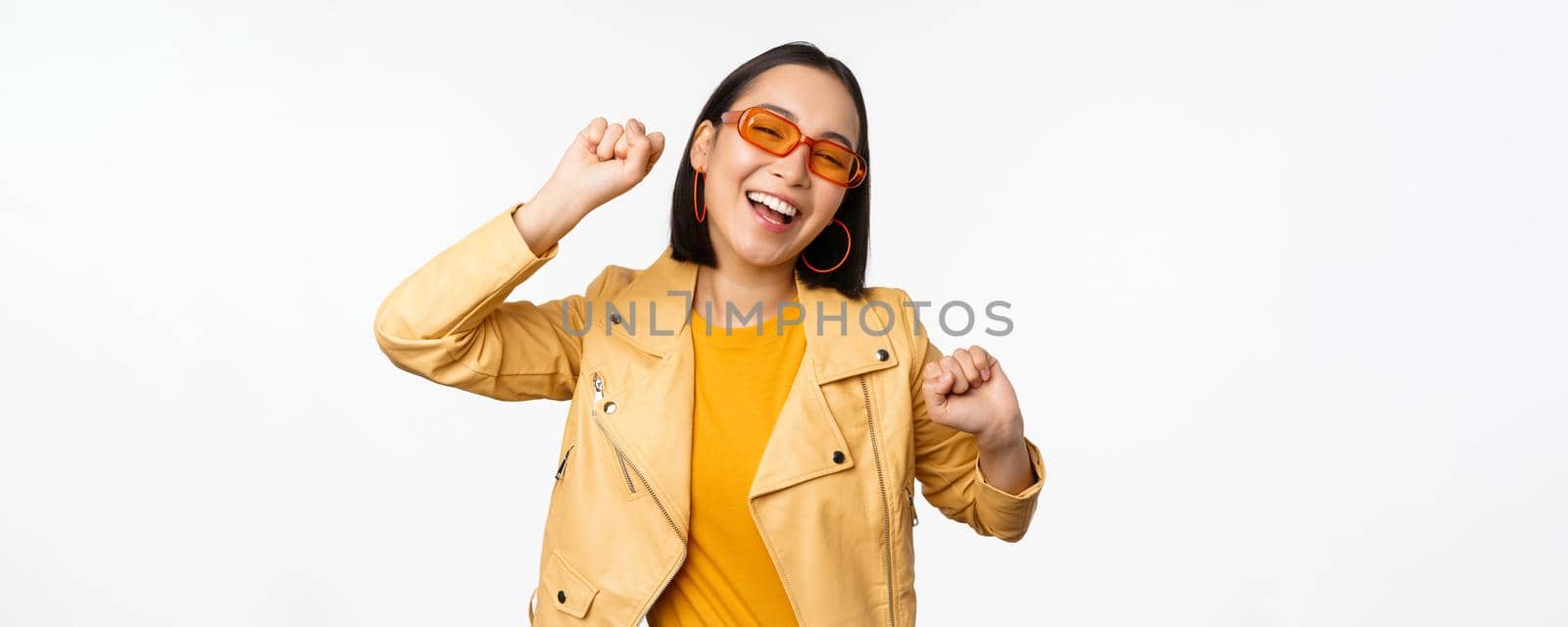 Happy stylish korean girl in sunglasses, dancing and laughing, smiling carefree, standing over white background. Copy space