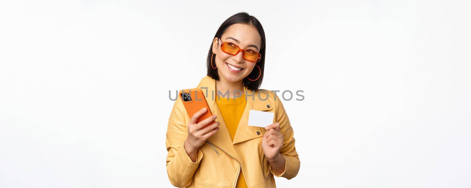 Online shopping and delivery concept. Happy korean girl in stylish clothes, holding credit card and smartphone, laughing and smiling, standing over white background.