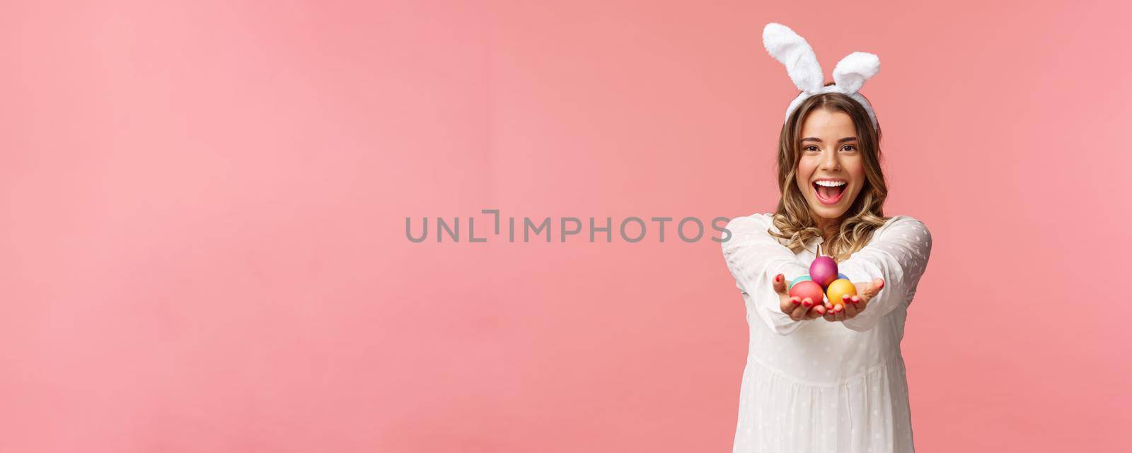 Holidays, spring and party concept. Portrait of lovely, romantic young blond woman in rabbit ears and white dress, giving you painted easter eggs as celebrating orthodox holiday, pink background.