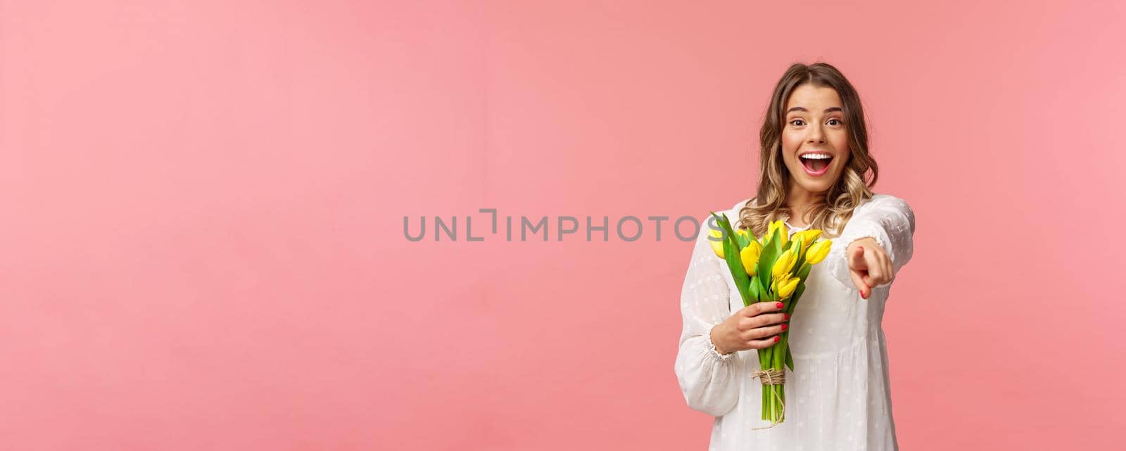 Holidays, beauty and spring concept. Portrait of excited happy and upbeat young blond girl in white dress, holding bouquet of yellow tulips, pointing finger at camera amazed, pink background by Benzoix