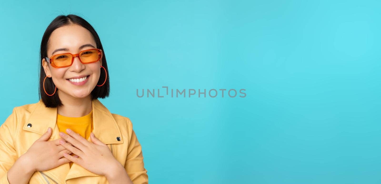 Close up portrait of asian young woman in sunglasses, smiling and looking romantic, standing happy over blue background. Copy space