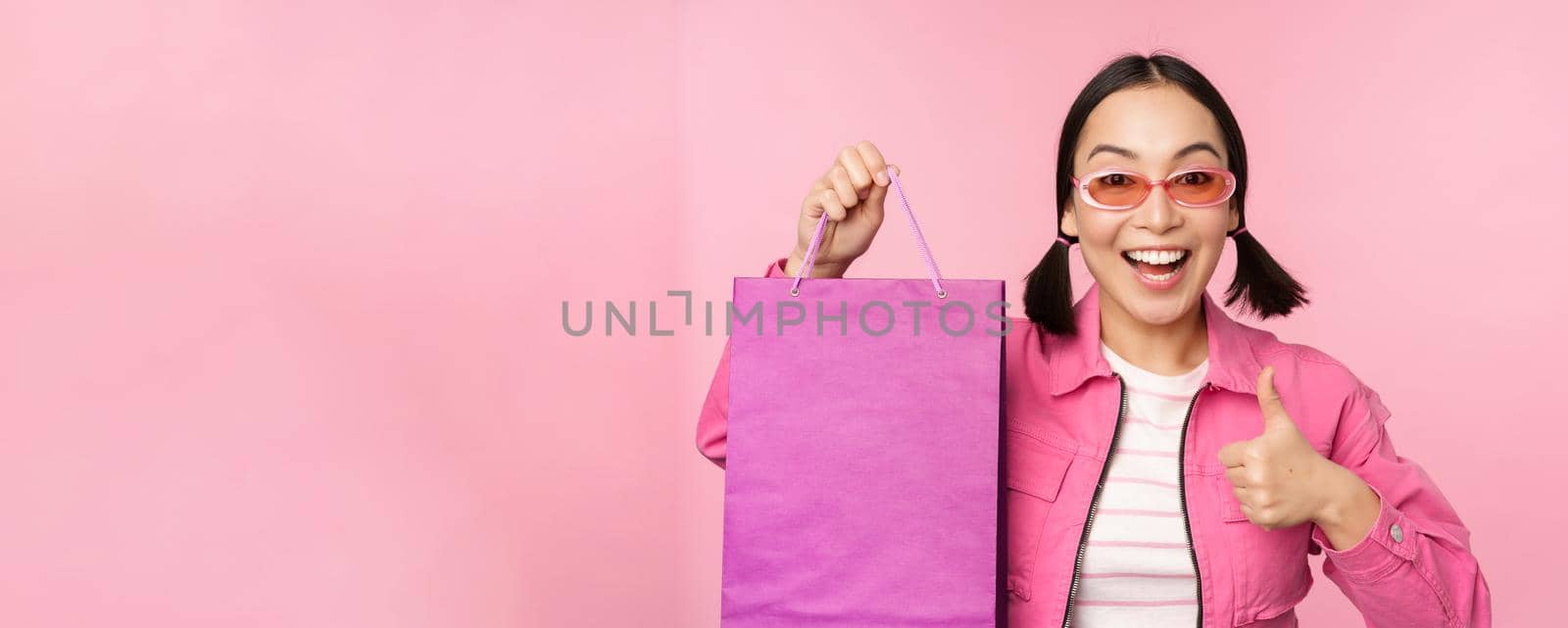 Shopping. Stylish asian girl in sunglasses, showing bag from shop and smiling, recommending sale promo in store, standing over pink background by Benzoix