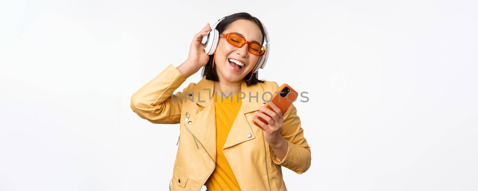 Technology concept. Stylish asian girl in headphones, holding smartphone, dancing and singing, listening music, standing over white background by Benzoix