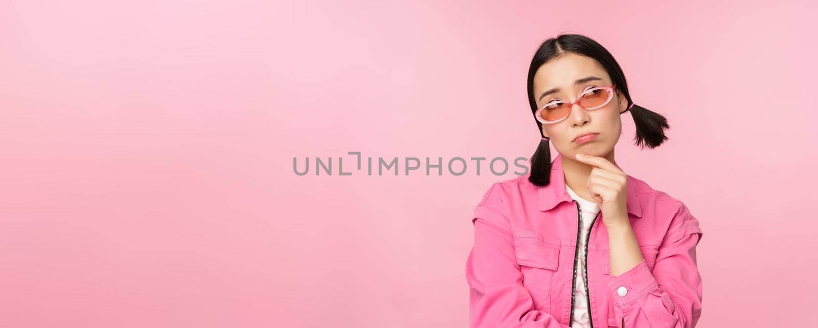 Portrait of sad and gloomy asian girl sulking from disappointment, standing upset against pink studio background by Benzoix