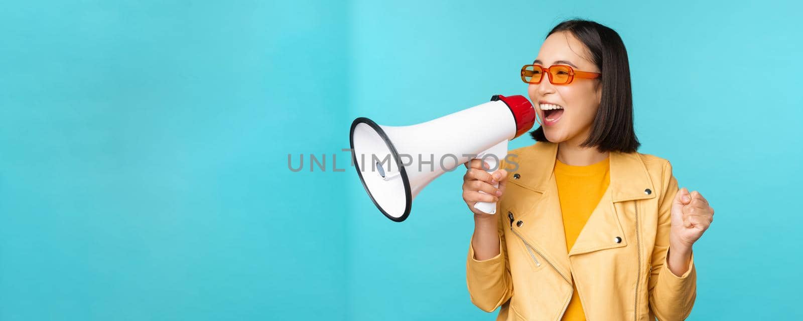 Young enthusiastic korean girl in trendy outfit, shouting in megaphone, making announcement, advertising, screaming in speakerphone, standing over blue background.