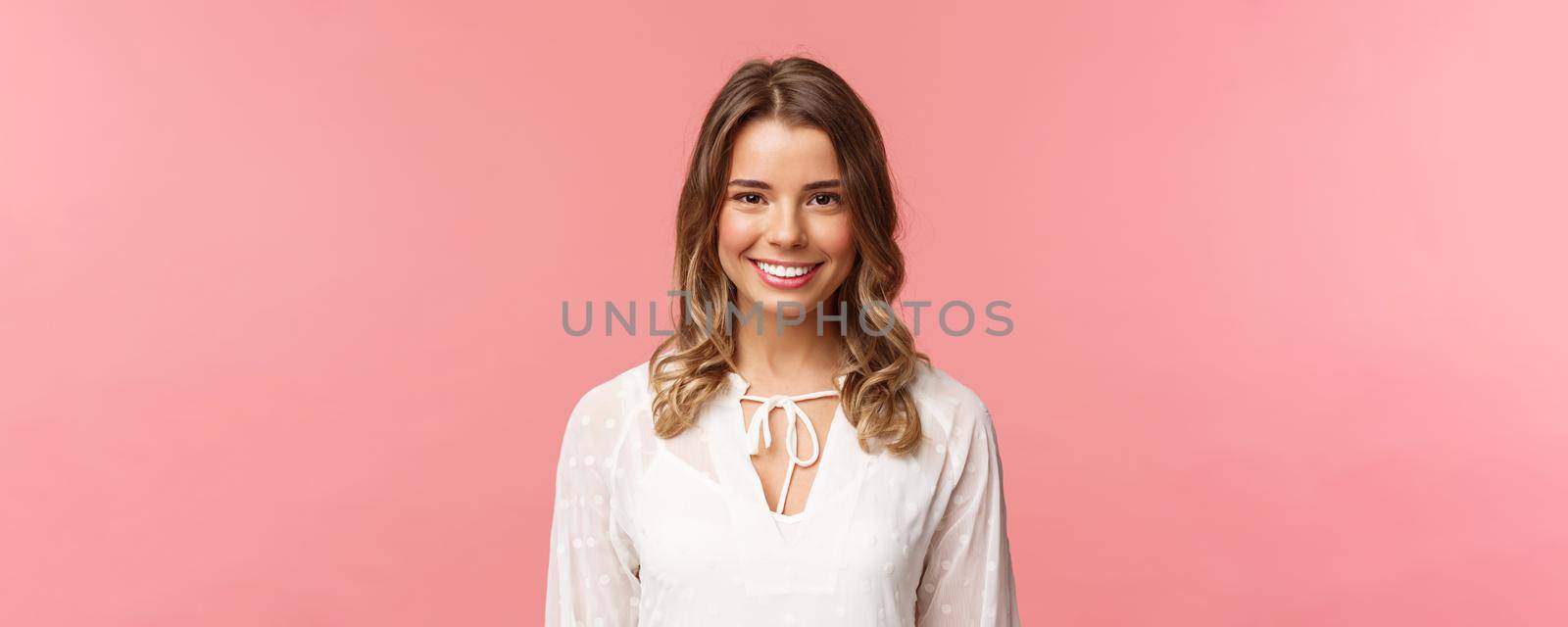 Close-up portrait of charming blond girl in lovely white dress with beaming smile looking at camera, feeling happy and cheerful, enjoying sunny romantic spring days, pink background.