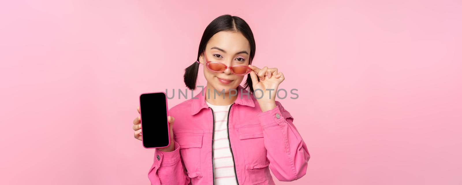 Stylish korean girl, young woman in sunglasses showing smartphone screen, mobile phone app interface or website, standing over pink background.