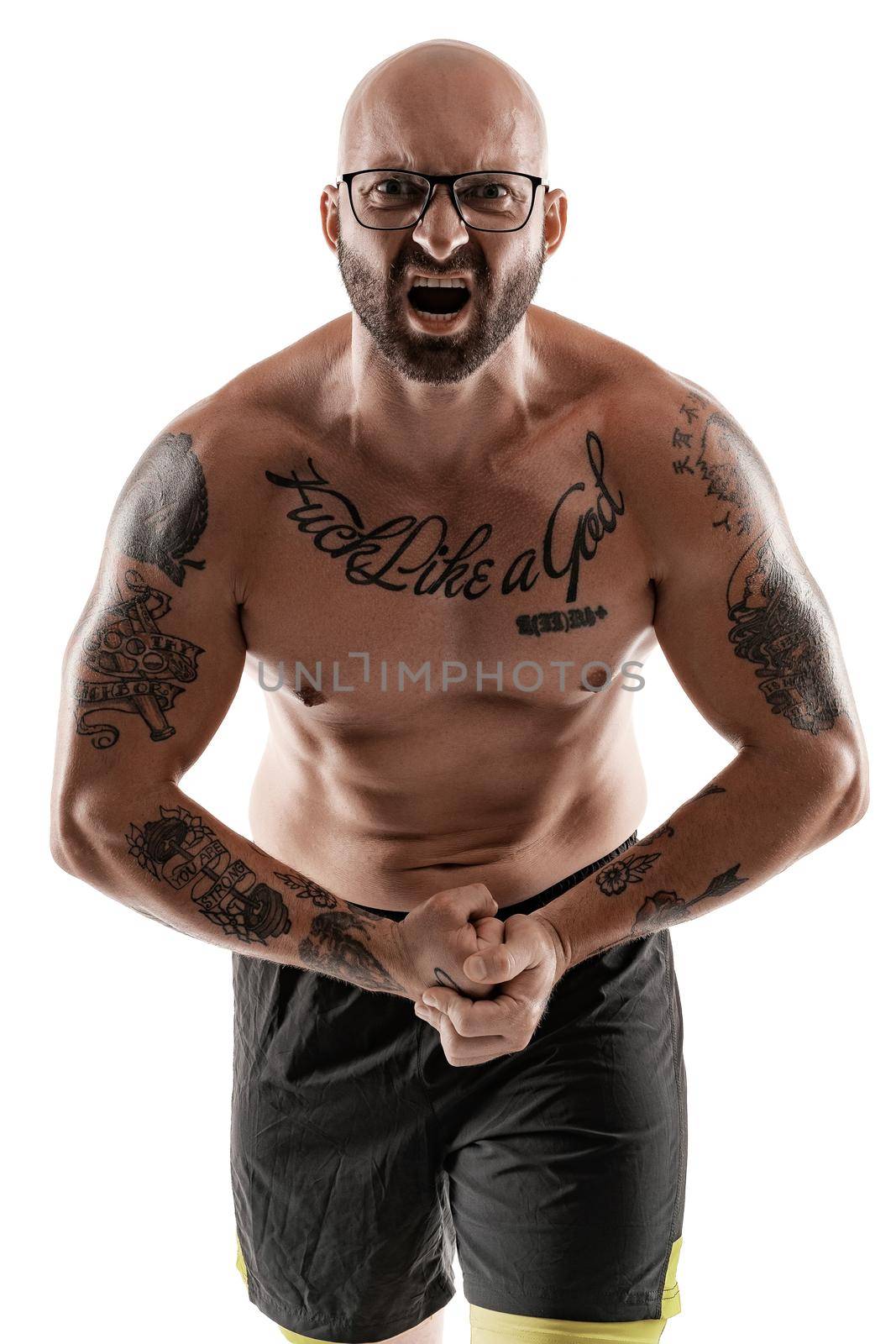 Handsome bald, bearded, tattooed man in glasses, black shorts is posing and showing his muscles isolated on white background, screaming and looking at the camera. Chic muscular body, fitness, gym, healthy lifestyle concept. Close-up portrait.