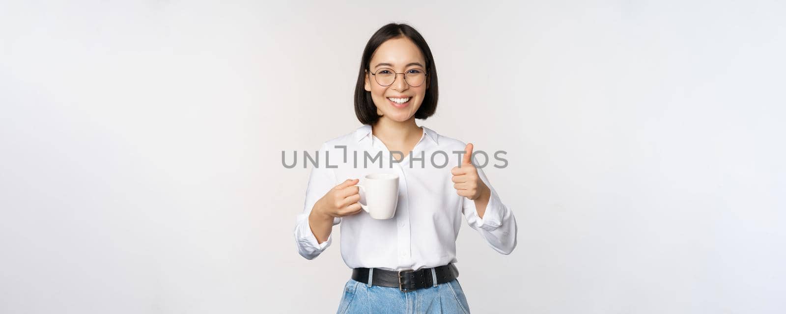 Smiling asian girl holding mug, white cup and thumbs up, recommending drink, coffee or tea, standing over white background by Benzoix