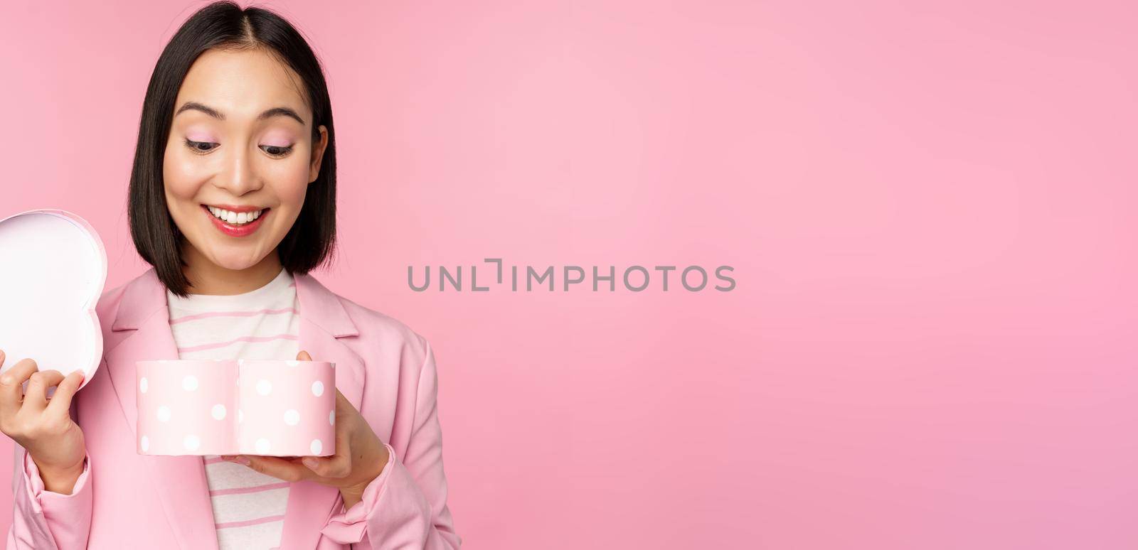 Happy cute korean girl in suit, opens up heart shaped box with romantic gift on white day holiday, standing in suit over pink background.