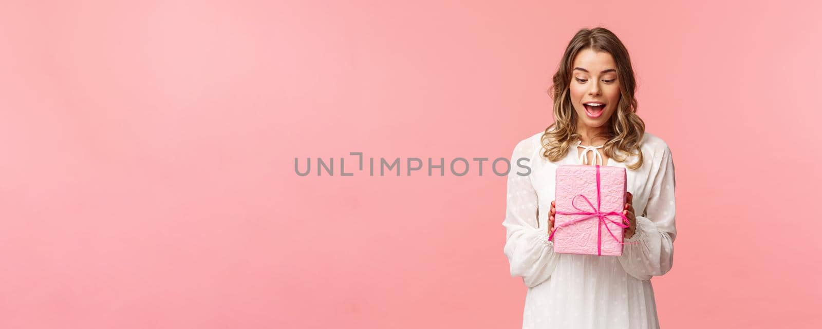 Holidays, celebration and women concept. Portrait of surprised charming young blond girl receive surprise gift, holding present in pink box look at it amused, curious whats inside, studio background by Benzoix