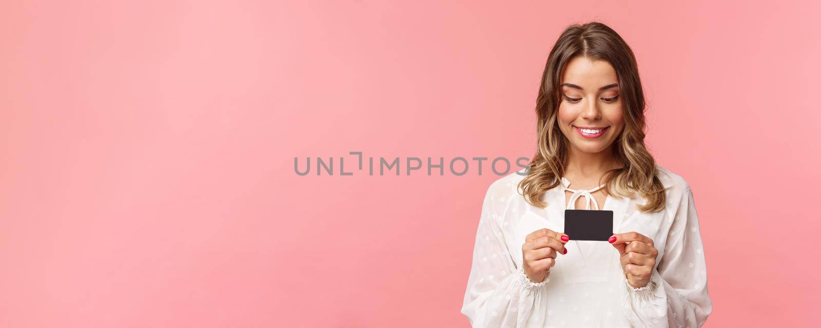 Close-up portrait of excited and amused blond girl in white dress, holding credit card and smiling thrilled, cant resist temptation to buy something, waste money online shopping, pink background.