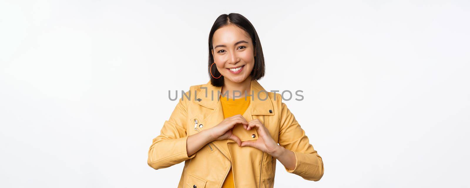 Image of beautiful asian girl showing heart, love sign, express care and romantic feelings, standing over white studio background by Benzoix