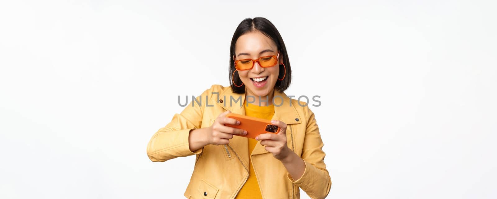Stylish korean girl in sunglasses, playing mobile video game, laughing and smiling while using smartphone, standing over white background by Benzoix