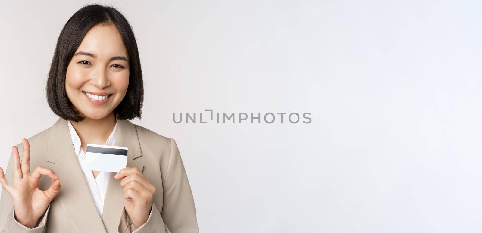 Smiling office clerk, asian corporate woman showing credit card and okay sign, recommending bank, standing over white background in beige suit.