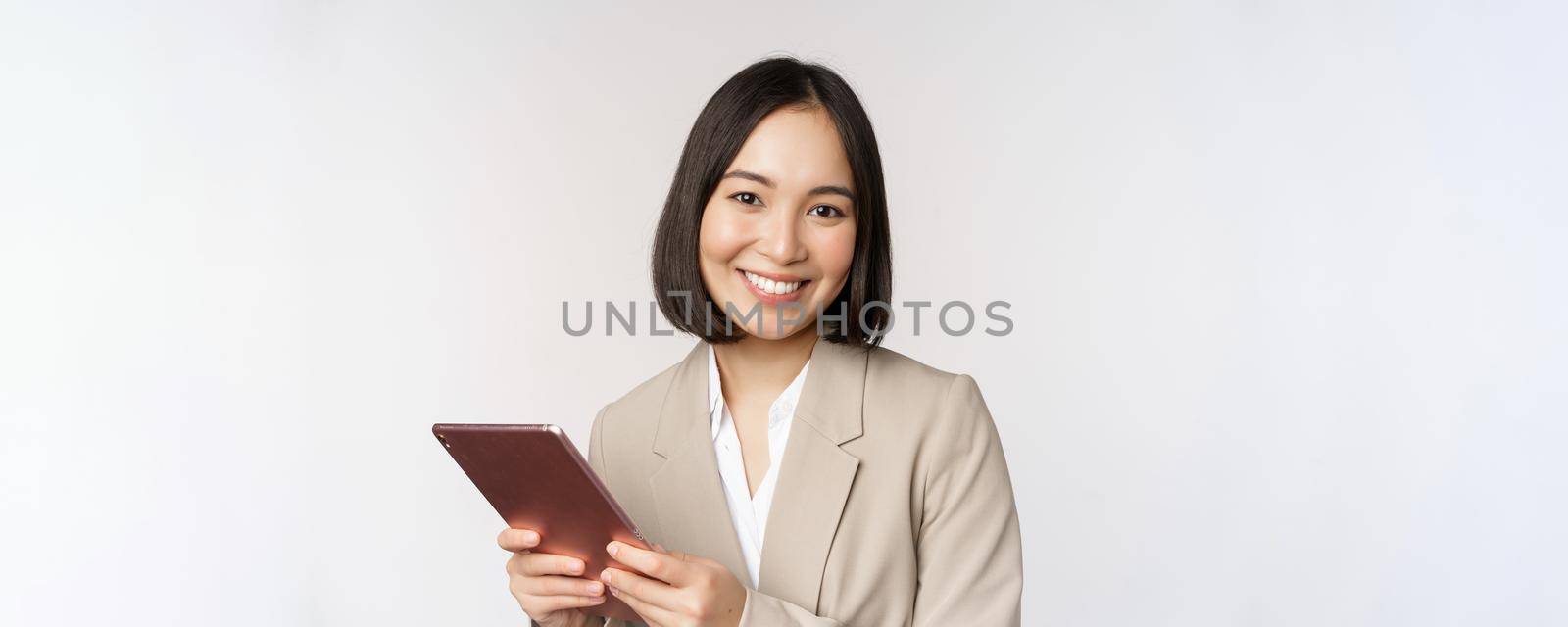 Image of asian businesswoman, saleswoman holding digital tablet and smiling, working with gadget, standing in suit over white background.