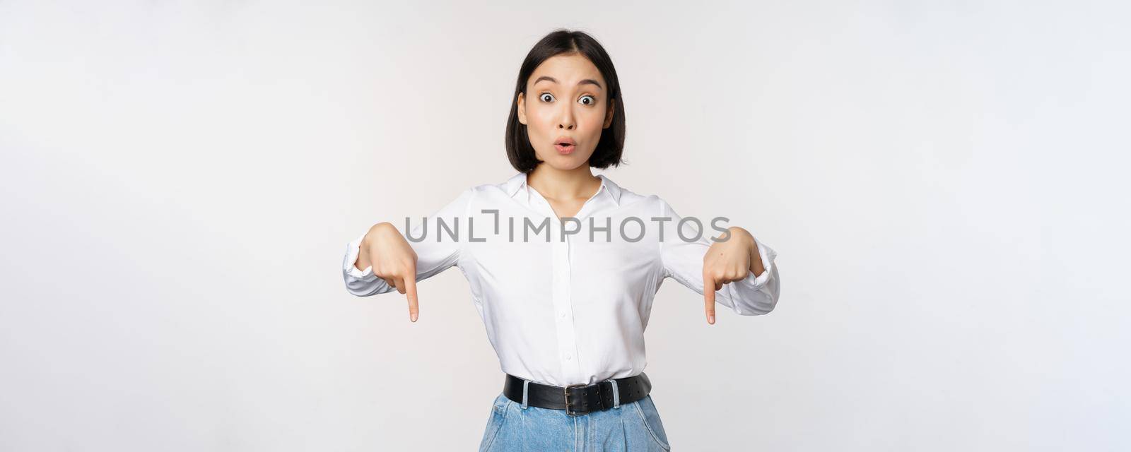 Portrait of young asian woman pointing fingers down and smiling, showing banner, click on link below gesture, inviting people to follow, standing over white background.