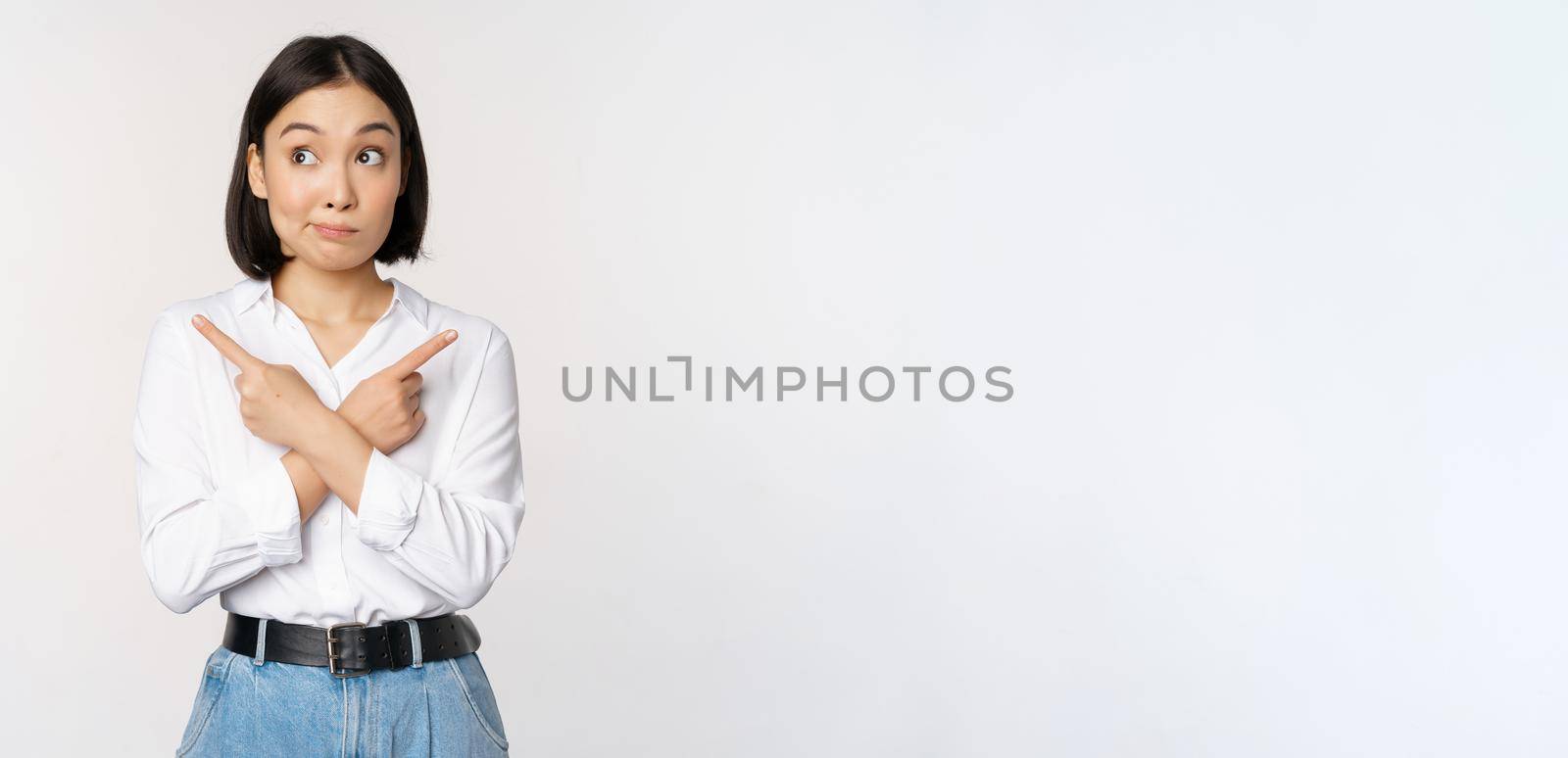 Indecisive asian woman pointing fingers sideways, pointing fingers and looking clueless, confused with choices, standing over white background.