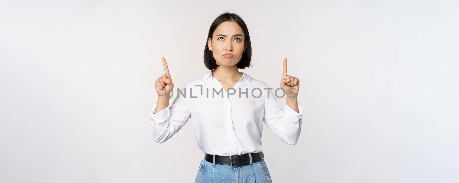 Skeptical businesswoman, asian office manager, pointing fingers up and grimacing doubtful, hesitating, standing over white background by Benzoix