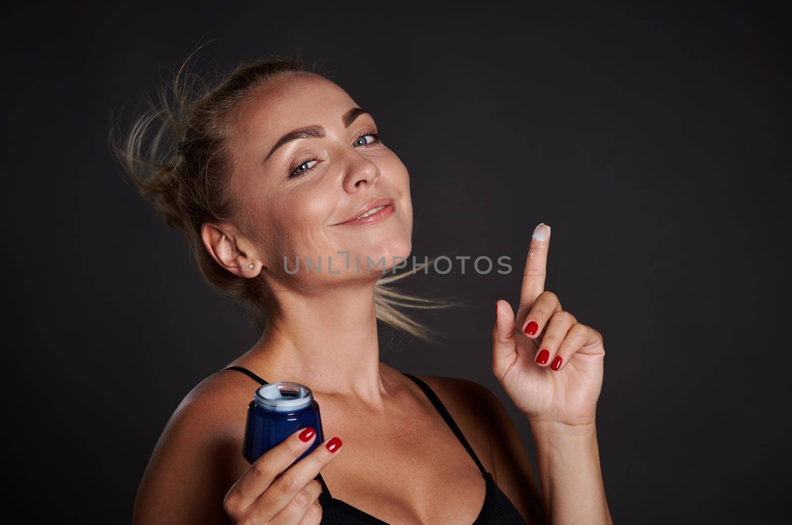 Beautiful middle aged woman smiles while holding anti wrinkles facial cream in her hands. Youth concept and prevention of first signs of aging process. Advertising studio shot over black background by artgf