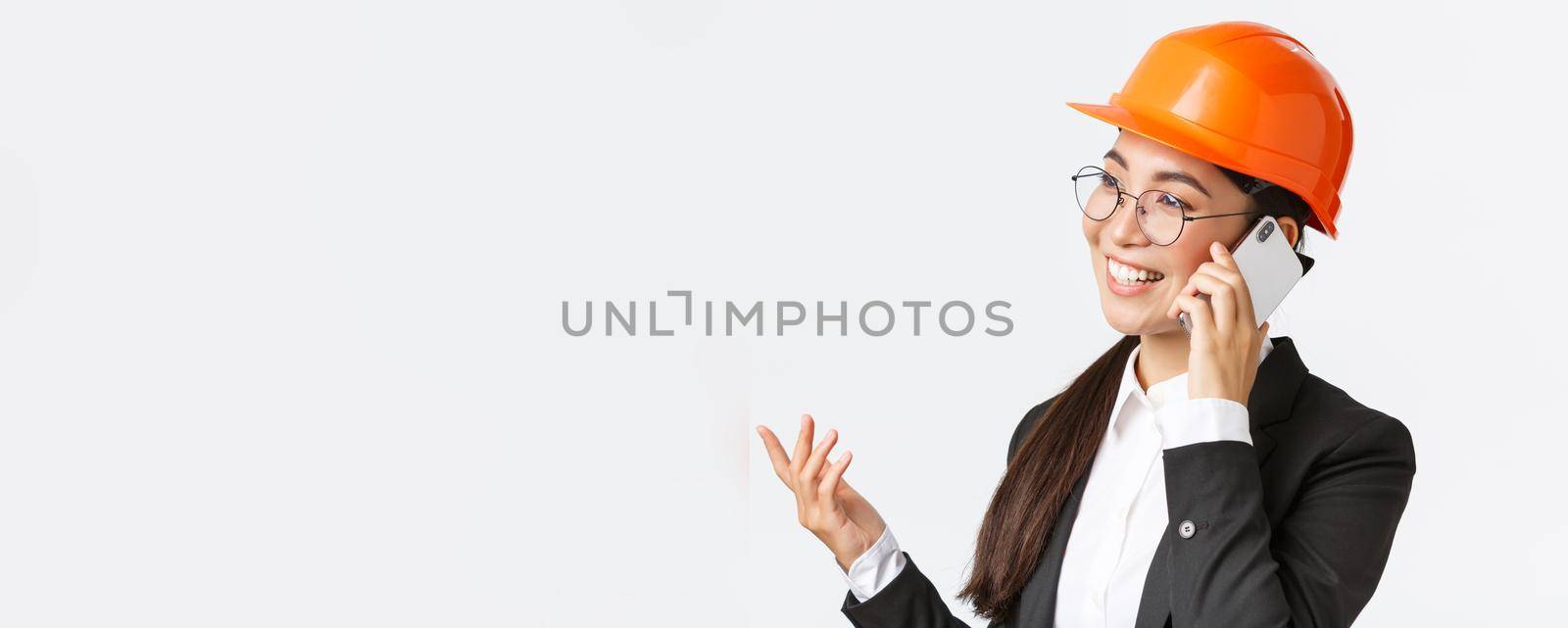 Close-up of professional smiling asian female entrepreneur at factory, chief engineer in safety helmet and suit, talking on phone, having business conversation with investors of enterprise.