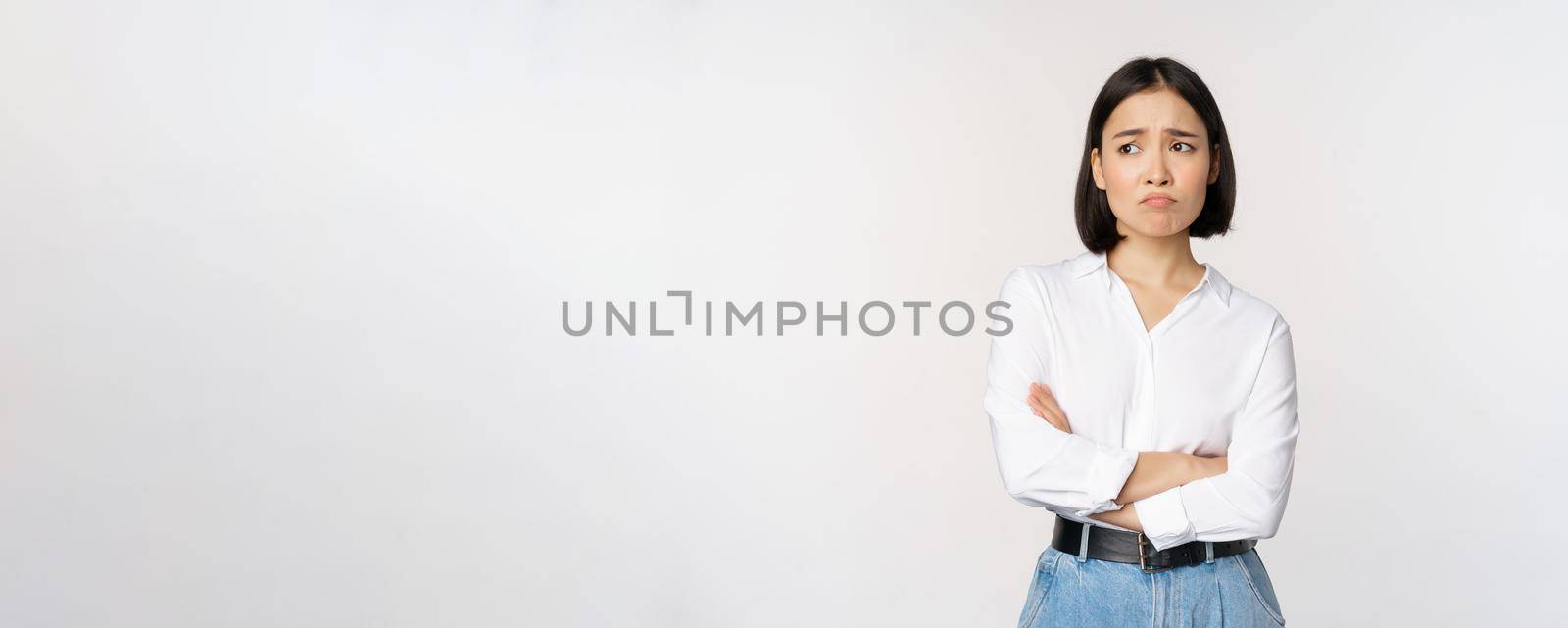 Image of sad office girl, asian woman sulking and frowning disappointed, standing upset and distressed against white background.