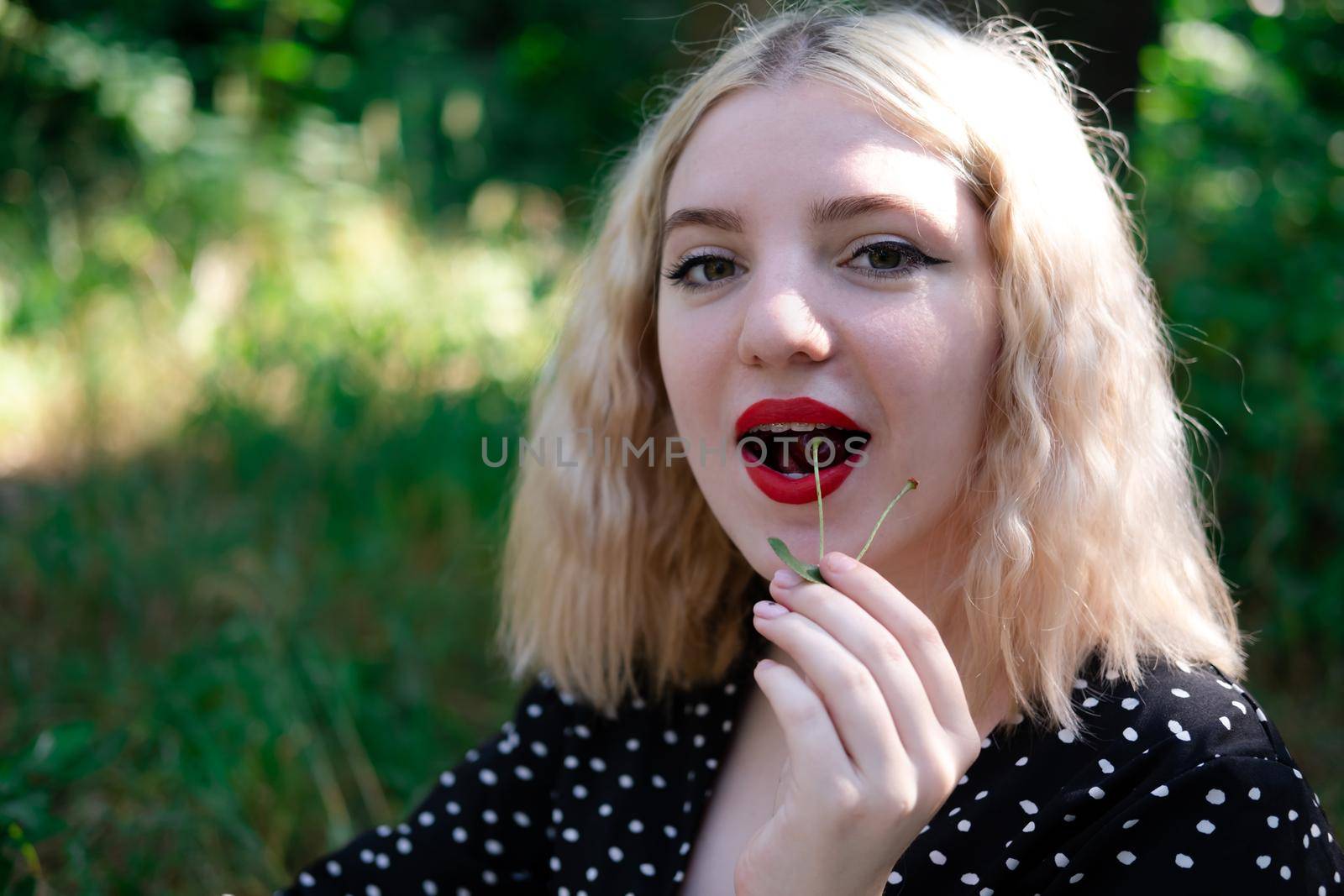 portrait of a charming blonde teenage girl wearing teeth braces biting a sweet cherry.