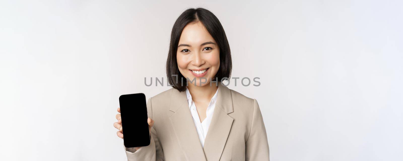 Portrait of corporate asian woman showing smartphone app interface, mobile phone screen, standing over white background.