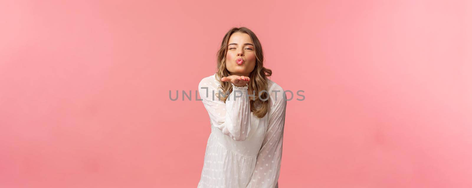 Beauty, fashion and women concept. Portrait of romantic tender, young blond woman in white trendy dress, sending air kiss at camera with sassy flirty smile, hold hand near lips, pink background.