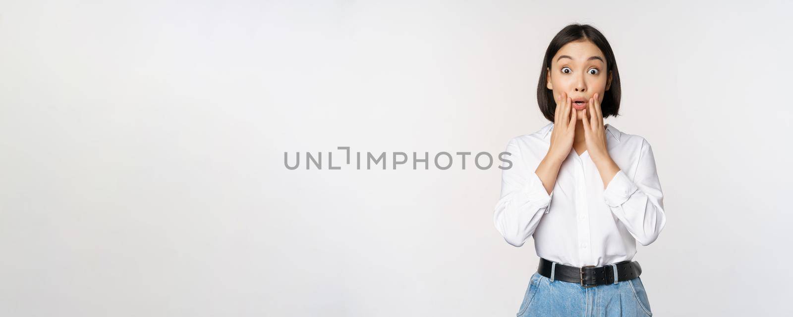 Portrait of surprised young office woman, asian businesswoman gasping amazed, saying wow, standing impressed of news against white background by Benzoix