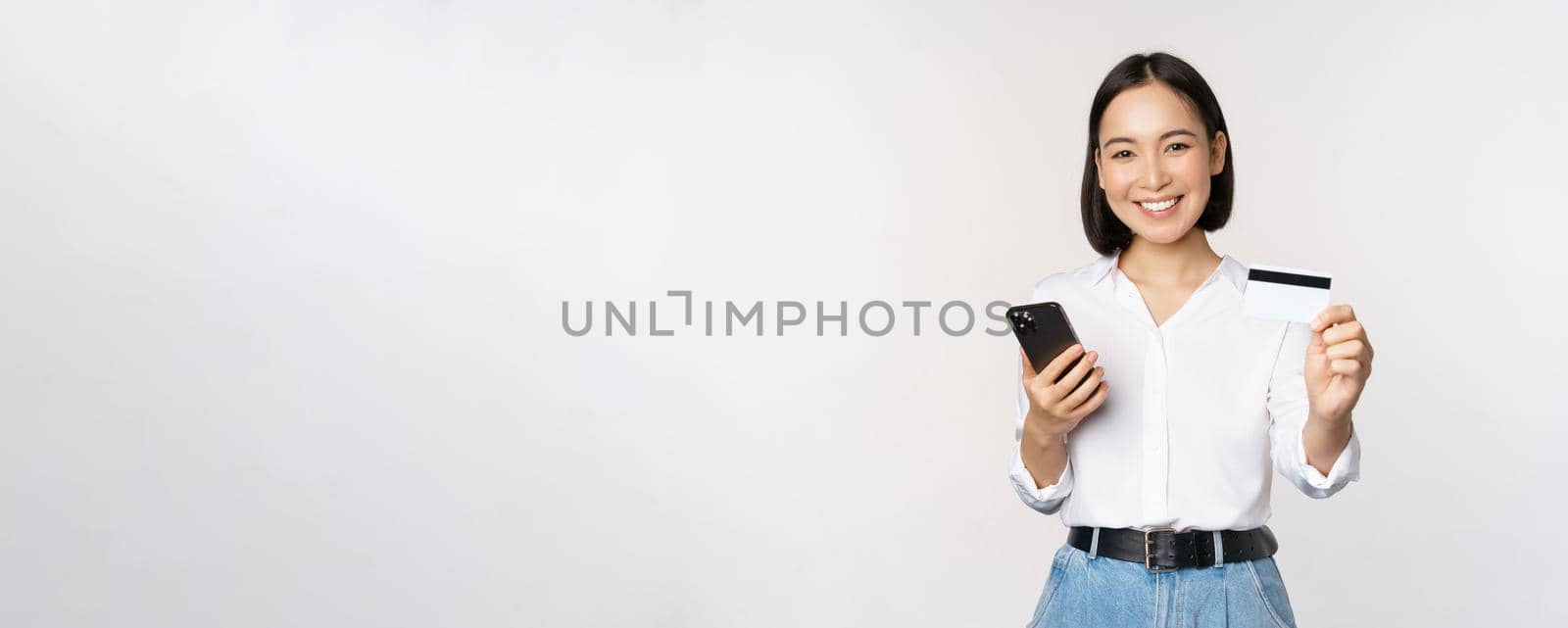 Online shopping concept. Smiling modern asian girl shows her credit card, holds mobile phone, order with smartphone, standing over white background.