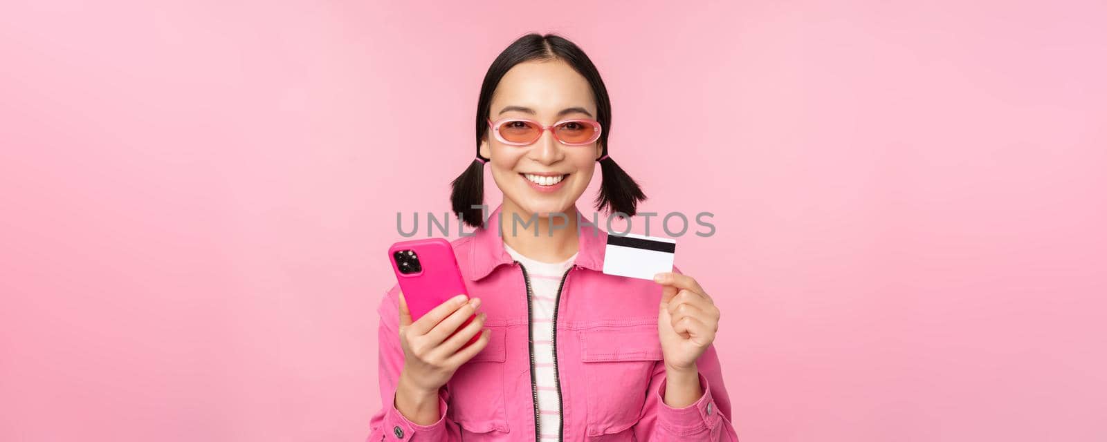 Online shopping. Smiling asian girl shopper, holding smartphone and credit card, paying in mobile app, standing over pink background.