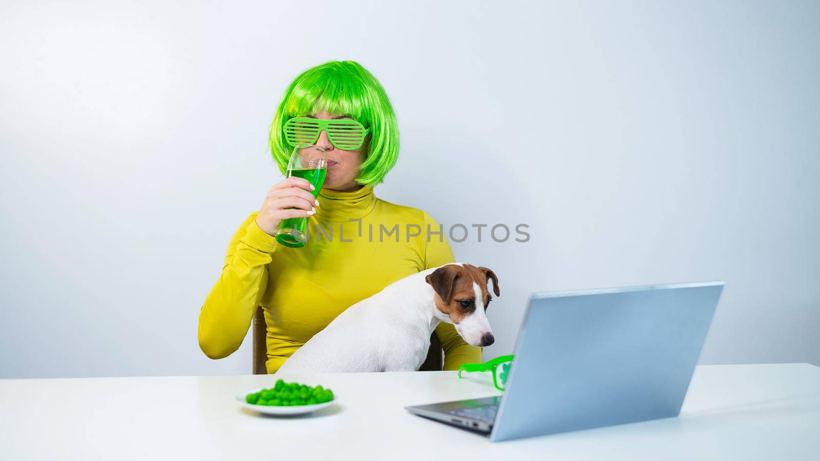 A young woman in a green wig and cheerful glasses drinks beer and bites glazed nuts. A girl sits with a dog at a table and celebrates st patrick's day online chatting with friends on a laptop. by mrwed54