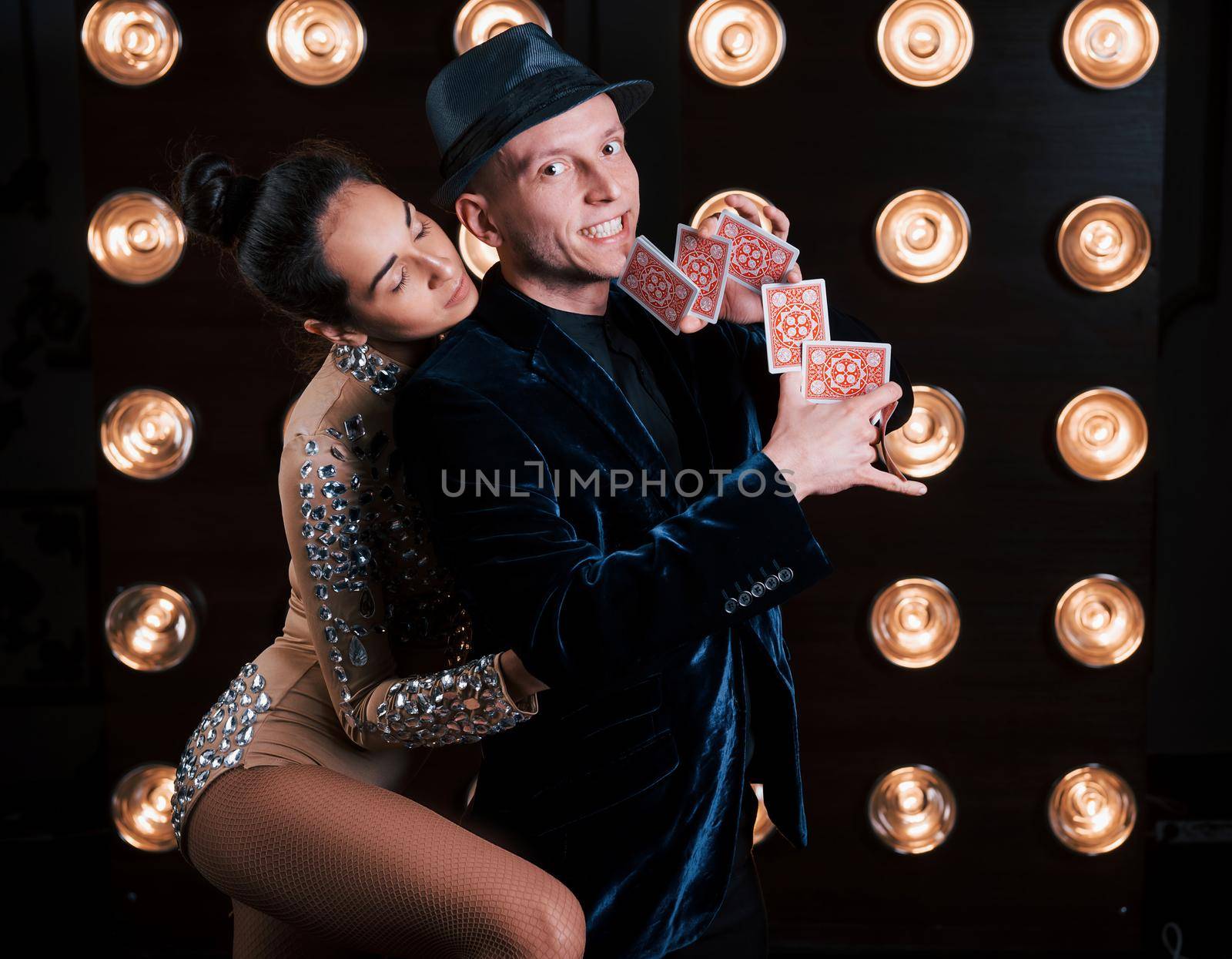 Girl is huggng her man while he showing tricks with cards. Happy couple posing for the portrait. Family business of magician tricks and shows.