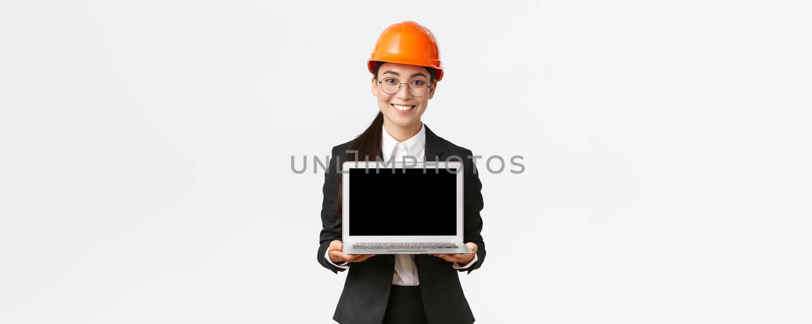 Smiling professional female asian engineer introduce construction plan to investors or clients, standing in safety helmet and suit showing laptop screen with pleased smile, white background.