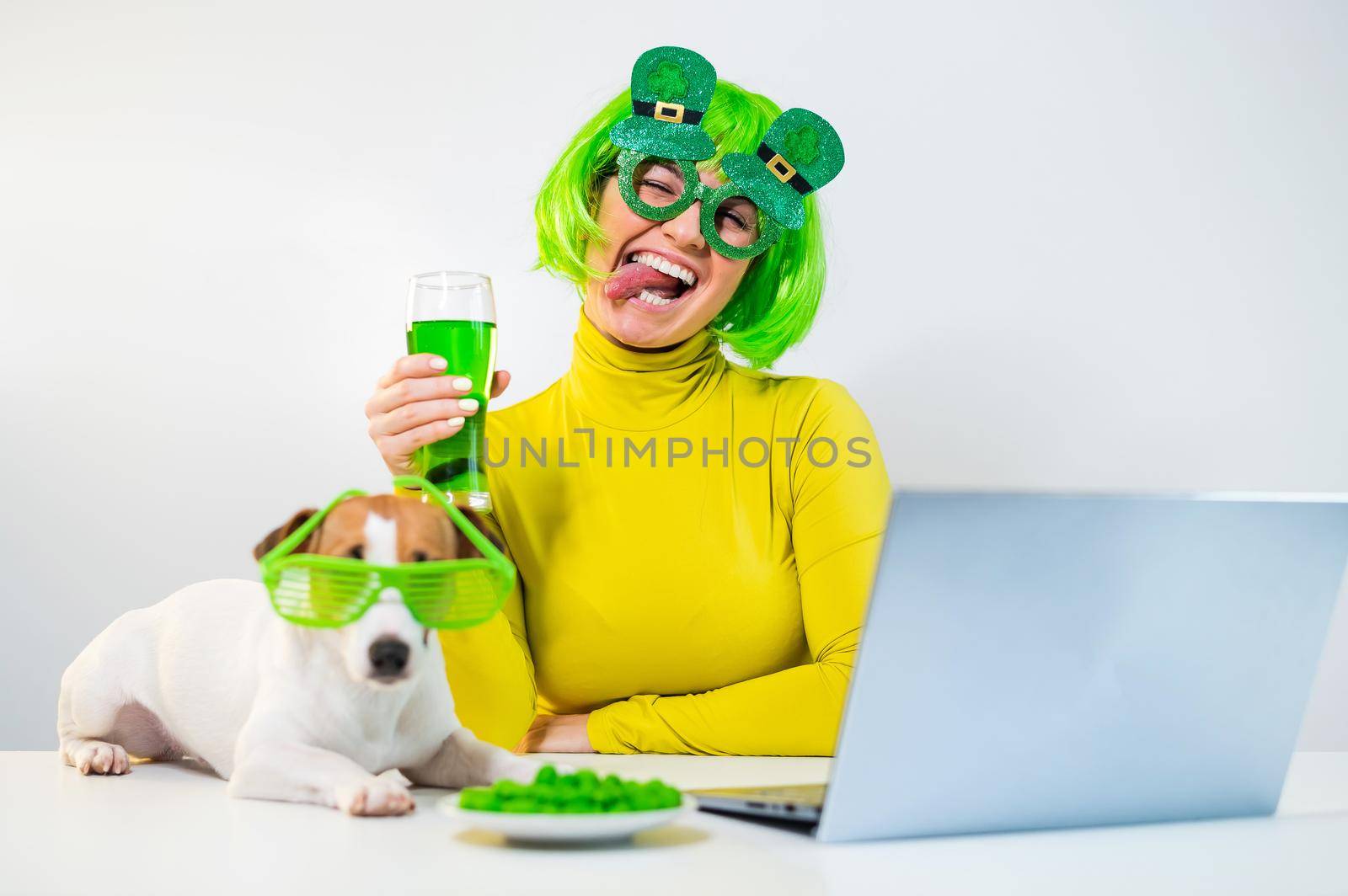 A young woman in a green wig and cheerful glasses drinks beer and bites glazed nuts. A girl sits with a dog at a table and celebrates st patrick's day online chatting with friends on a laptop. by mrwed54