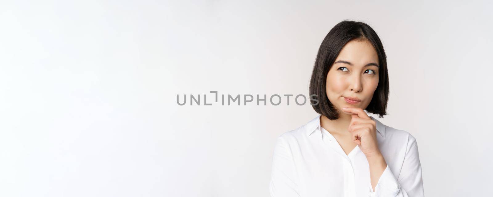 Close up portrait of asian woman thinking, looking aside and pondering, making decision, standing over white background.