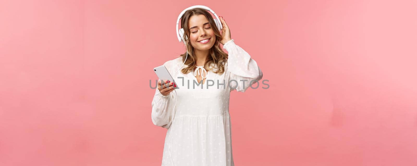 Portrait of tender happy and cute blond girl in white dress, close eyes and smiling pleased as listening music in headphones, feeling warmth and happiness enjoying favorite track, hold mobile phone.