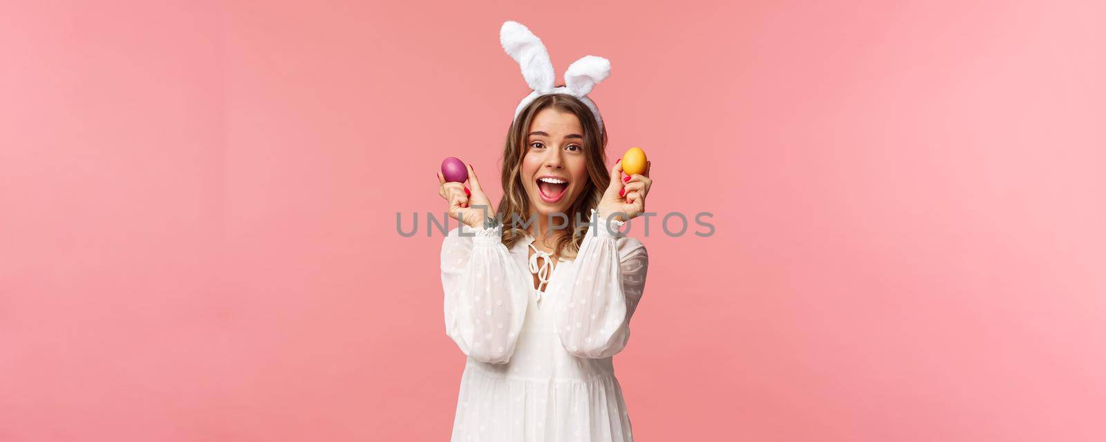 Holidays, spring and party concept. Portrait of excited charming young woman celebrating Easter in rabbit ears and white party dress, dancing with two painted eggs, pink background by Benzoix