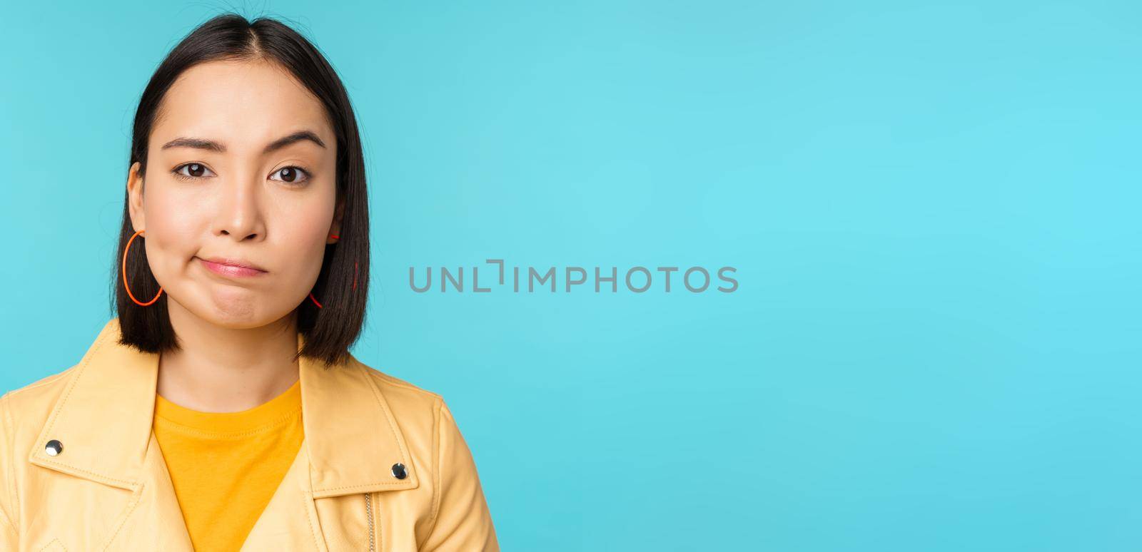 Close up of skeptical asian girl looking disappointed, staring with doubt or disbelief, grimacing at camera, standing over blue background.