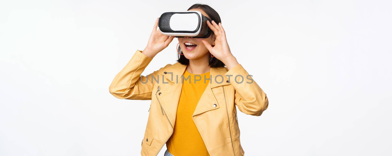 Enthusiastic asian girl in VR glasses, looking with virtual reality headset at smth amazing, standing over white background. Copy space