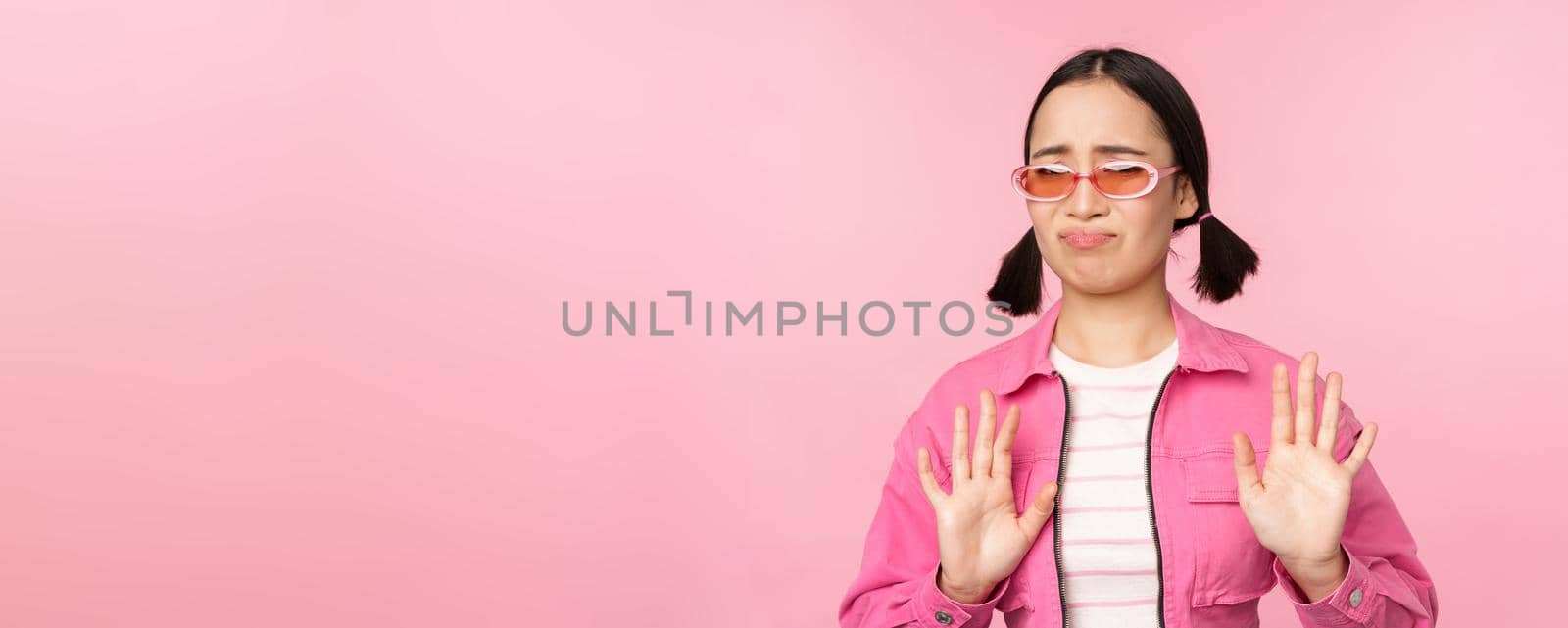 No thanks. Disgusted asian girl in sunglasses, rejecting smth, shaking hands to refuse, decline, standing over pink background.