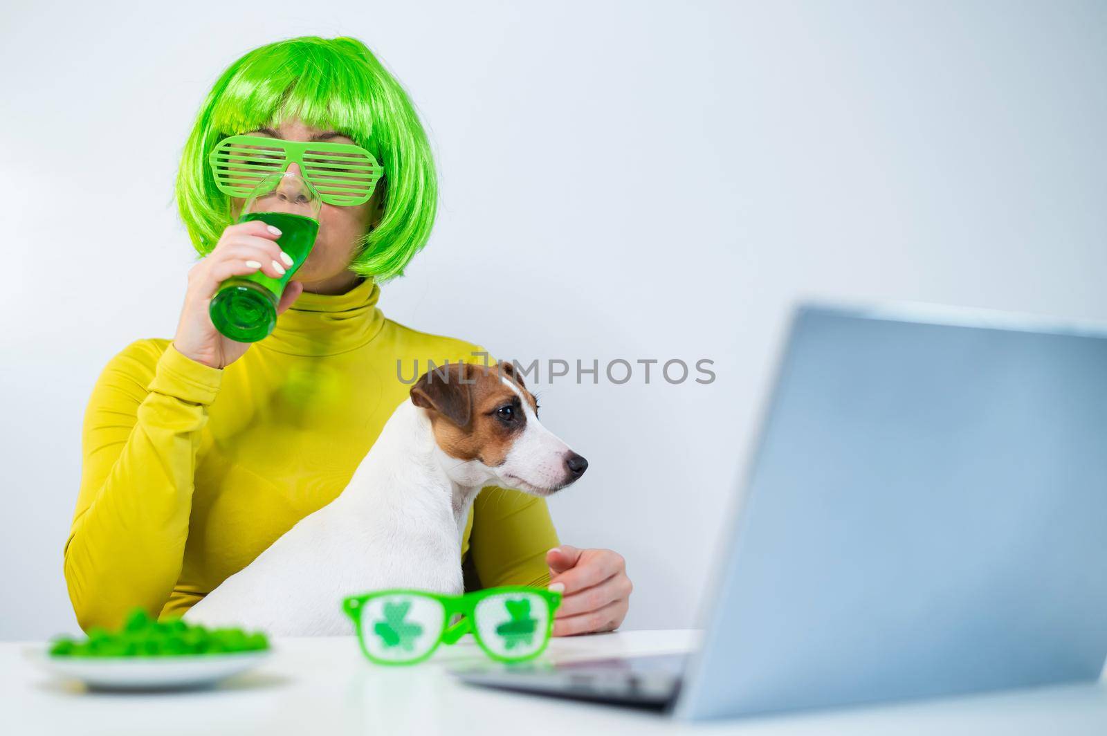 A young woman in a green wig and cheerful glasses drinks beer and bites glazed nuts. A girl sits with a dog at a table and celebrates st patrick's day online chatting with friends on a laptop