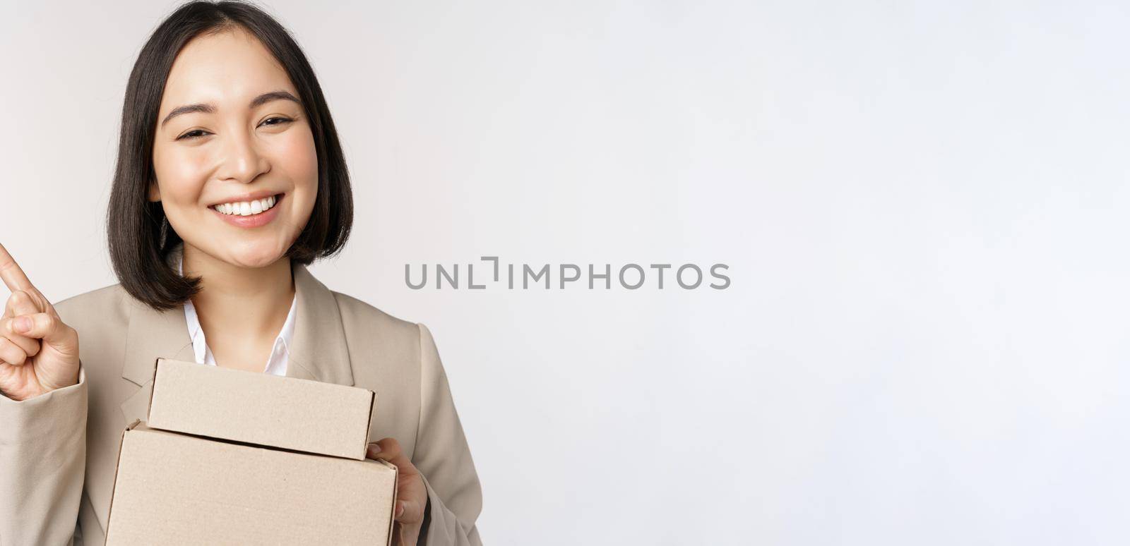 Smiling asian saleswoman pointing finger at copy space, holding boxes with delivery order, standing over white background by Benzoix
